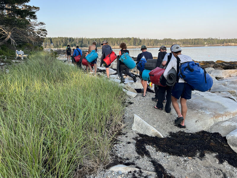 A group of people are walking along a rocky shoreline, carrying large backpacks and sleeping bags. They appear to be on a camping or hiking trip, possibly heading to or from a campsite. The scene is set near the coast, with a body of water visible in the background and greenery on the side. The weather seems fair. 